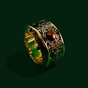 May include: A gold ring featuring an intricate black and gold design, with a dark red gemstone at its centre.