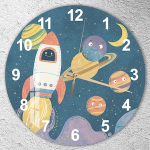 Op de afbeelding: Een ronde wandklok met een ruimtethema. De wijzerplaat is donkerblauw met een raket, planeten en sterren. De cijfers zijn wit en de wijzers goudkleurig. De raket is wit en oranje, met een lachend gezicht.