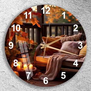 May include: A round wall clock featuring a printed image of a cozy porch scene with autumn leaves, a bench with pillows and a throw blanket, and glowing candles. The clock face has white numbers from 1 to 12 and thin gold-colored metal hands.