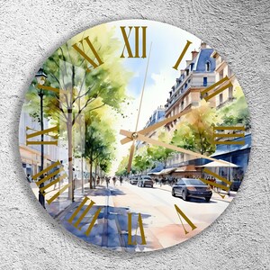 Pode incluir: Um relógio redondo com uma pintura em aquarela de uma cena de rua parisiense. O mostrador do relógio tem algarismos romanos e ponteiros dourados. A pintura apresenta edifícios, árvores e pessoas caminhando pela rua.