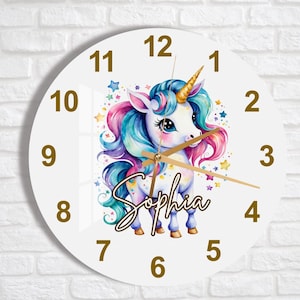 Puede incluir: Reloj redondo blanco con números y manecillas dorados. Un unicornio colorido con un cuerno dorado y una crin arcoíris está en el centro del reloj. El nombre del unicornio, "Sophia", está escrito en cursiva dorada debajo del unicornio.