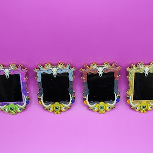 May include: Four decorative, rectangular mirrors with ornate gold frames. Each mirror features a black reflective surface and is adorned with a skull and eye-shaped embellishments in various colors: purple, blue, red, and yellow. The mirrors are set against a vibrant purple background.