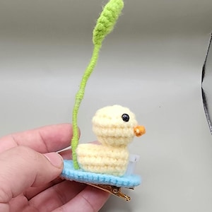 Könnte beinhalten: Handgefertigte Haarspange mit Häkelente. Die Ente ist gelb mit einem kleinen orangefarbenen Schnabel und schwarzen Augen und sitzt auf einer hellblauen Basis. Ein grüner Stiel mit einer grünen Spitze erstreckt sich nach oben. Die Spange ist goldfarben.