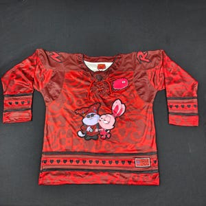 May include: A red long-sleeve jersey featuring a dark red swirling pattern, a lace-up neckline, and a graphic of two cartoon characters. The sleeves and hem have black stripes with heart motifs, and a small rectangular patch is on the lower front.