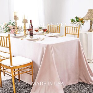 Può includere: Un tavolo da pranzo formale apparecchiato per un'occasione speciale. Il tavolo è coperto da una tovaglia di velluto rosa chiaro. Sedie, piatti e posate dorate sono disposti intorno al tavolo. Il testo "MADE IN USA" è visibile.