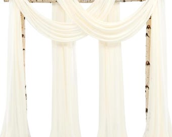 May include: A cream-colored wedding arch with draped fabric. The arch is constructed from birch branches and features flowing, sheer fabric. The fabric is arranged in a crisscross pattern across the top and hangs down in long panels.
