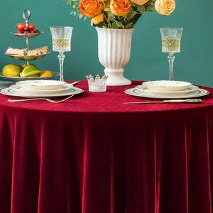 Pode incluir: Uma mesa de jantar formal com uma toalha de mesa de veludo vermelho escuro. A mesa está posta com porcelana fina, cristais e talheres dourados. Um vaso branco contém rosas laranja e amarelas. Uma bandeja de servir em camadas contém frutas e doces.