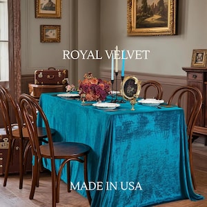 May include: A teal velvet tablecloth covers a dining table set with white plates and gold-coloured candle holders. A floral centrepiece and the text "ROYAL VELVET" and "MADE IN USA" are visible. The table is surrounded by wooden chairs.