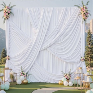 以下が含まれることがあります： 結婚式やイベント用の白いドレープ生地の背景で、ピンクと白の花のアレンジメントで飾られています。 金と白の風船と装飾的なテーブルが前景にあり、緑の芝生に置かれています。