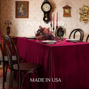 May include: A dining table draped with a deep red velvet tablecloth. The table is set with plates, glasses, and a floral arrangement. The wall features antique clocks and a framed painting. The text "ROYAL VELVET" and "MADE IN USA" are visible.