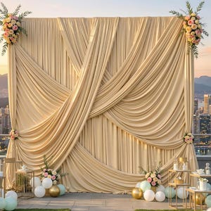May include: A large, draped backdrop in a warm champagne color, adorned with floral arrangements at the corners and base. The backdrop is set against a cityscape, with decorative tables and balloons in the foreground, creating an elegant event setting.