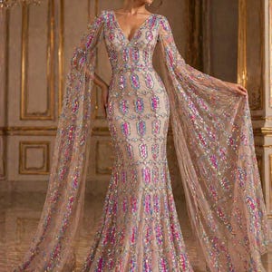 Puede incluir: Un vestido de noche beige, largo hasta el suelo, con escote pronunciado y mangas fluidas. El vestido está adornado con lentejuelas de colores en un patrón repetitivo. Las mangas son transparentes y se extienden desde los hombros, creando un efecto dramático. Adecuado para ocasiones especiales.
