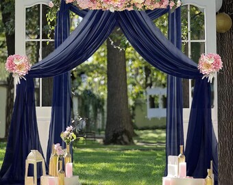 Può includere: Uno sfondo per una cerimonia di matrimonio con una cornice di porta bianca decorata con tessuto blu navy trasparente e una ghirlanda di fiori rosa e bianchi.
