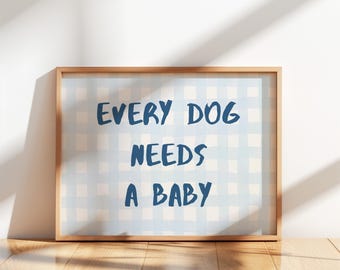 May include: A framed print with the text "EVERY DOG NEEDS A BABY" in navy blue on a light blue and white checkered background. The frame is a light brown color, and the print is displayed on a wooden surface.