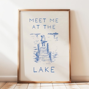 Puede incluir: Una impresión enmarcada con un fondo beige claro e ilustración azul. La imagen representa un perro saltando de un muelle a un lago. El texto "MEET ME AT THE LAKE" está impreso en azul arriba y abajo del perro.