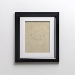 Op de afbeelding: Een zwart ingelijst print met een beige achtergrond en de tekst "it is so good to be home" geschreven in zwarte cursieve letters.