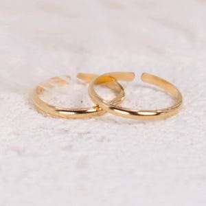 May include: Two gold-colored toe rings are displayed on a white, textured surface. The rings are open-ended and have a simple, elegant design. The rings are likely made of a precious metal.