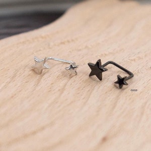 May include: Two ear cuffs, one silver-toned and one black, each featuring two star-shaped charms connected by a curved metal bar. The star charms vary in size, with a larger star at one end and a smaller star at the other end of the curved bar.