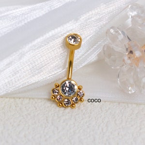 May include: Gold belly button ring with a clear crystal center and a circle of smaller clear crystals around it.  The ring is on a white fabric background.  The text "COCO" is visible in the lower right corner of the image.