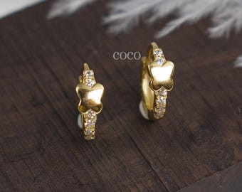 May include: A pair of gold hoop earrings featuring a small butterfly charm on each hoop. The hoops are embellished with small, clear, sparkling stones along the front surface. The word coco is printed in small black letters between the earrings.
