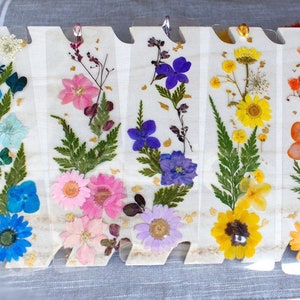 May include: Set of seven pressed flower bookmarks, each featuring a different arrangement of dried flowers and leaves. The bookmarks are clear and have a white background. The flowers are in various colors, including blue, pink, yellow, orange, and white.