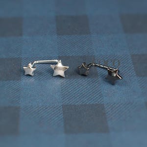 Puede incluir: Dos piercings de barra de metal en tono plateado, cada uno con dos extremos en forma de estrella. Las piezas de joyer&iacute;a est&aacute;n colocadas horizontalmente, mostrando un acabado liso y pulido en los motivos de estrella y las barras rectas.