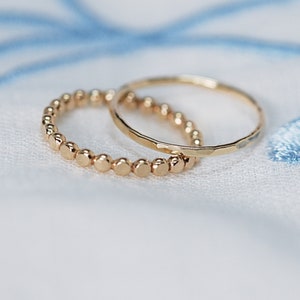 May include: Two gold rings on a white fabric background. One ring is a simple band, the other is a beaded ring.