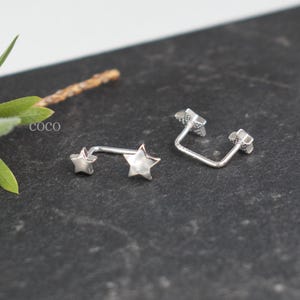 Puede incluir: Dos pendientes de metal plateado, cada uno con una estrella peque&ntilde;a en un extremo y una estrella m&aacute;s grande en el otro, unidos por una barra fina y curva. La palabra coco est&aacute; impresa en letras blancas peque&ntilde;as cerca de las piezas.