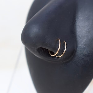 May include: Two gold nose rings on a black nose piercing model. The rings are thin and delicate, and they are stacked on top of each other.