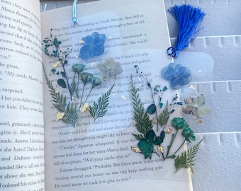 Puede incluir: Un marcapáginas con flores prensadas azules y verdes y un borla azul. El marcapáginas está en una página de un libro con texto sobre una familia y sus relaciones. El texto en la página dice "Yep, they did. According to Uncle Mason, they fell in the flea market while digging through a box of old girls' cards."