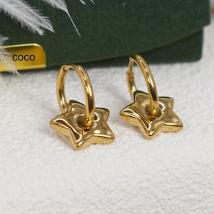 May include: A pair of gold-colored hoop earrings with star-shaped charms. The earrings are displayed on a white surface, with a green background and a small gold label that reads "coco". The earrings are a bright, shiny gold color.