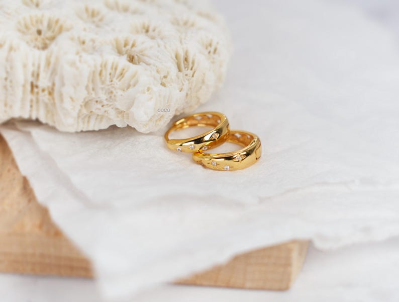 May include: A pair of gold hoop earrings featuring a polished finish and small, embedded star-shaped accents with clear stones. The earrings have a hinged closure mechanism and a rounded profile, showcasing a bright metallic yellow gold tone.