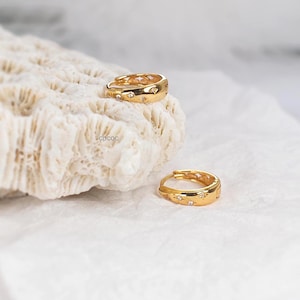May include: A pair of gold hoop earrings featuring small, embedded clear stones arranged in a star-like pattern. One earring rests on a textured white surface, while the other is positioned slightly below it. The word COCOC is printed in small black letters.