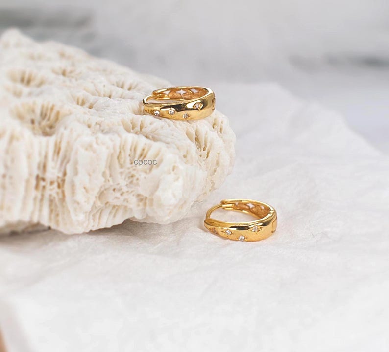 May include: A pair of gold hoop earrings featuring small, embedded clear stones arranged in a star-like pattern. One earring rests on a textured white surface, while the other is positioned slightly below it. The word COCOC is printed in small black letters.