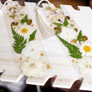 May include: Two white pressed flower bookmarks with white tassels. The bookmarks are made of clear acrylic and feature pressed white flowers, green ferns, and gold accents.