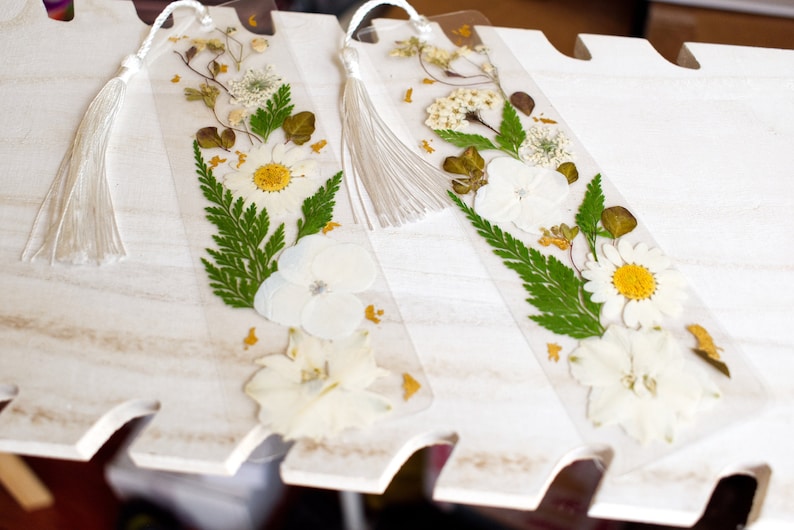 May include: Two white pressed flower bookmarks with white tassels. The bookmarks are made of clear acrylic and feature pressed white flowers, green ferns, and gold accents.