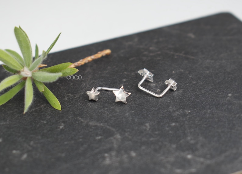 Puede incluir: Dos pendientes de metal plateado, cada uno con una estrella peque&ntilde;a en un extremo y una estrella m&aacute;s grande en el otro, unidos por una barra fina y curva. La palabra coco est&aacute; impresa en letras blancas peque&ntilde;as cerca de las piezas.