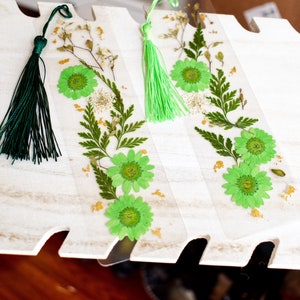 May include: Three green and white pressed flower bookmarks with green tassels. The bookmarks are on a white wooden surface.