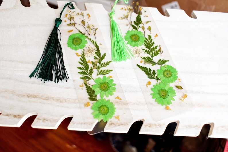 May include: Three green and white pressed flower bookmarks with green tassels. The bookmarks are on a white wooden surface.
