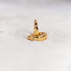 May include: A pair of gold hoop earrings featuring a polished finish and small, star-shaped engravings with embedded clear stones. The word coco is printed in small black letters to the left of the jewellery. The hoops have a hinged closure.