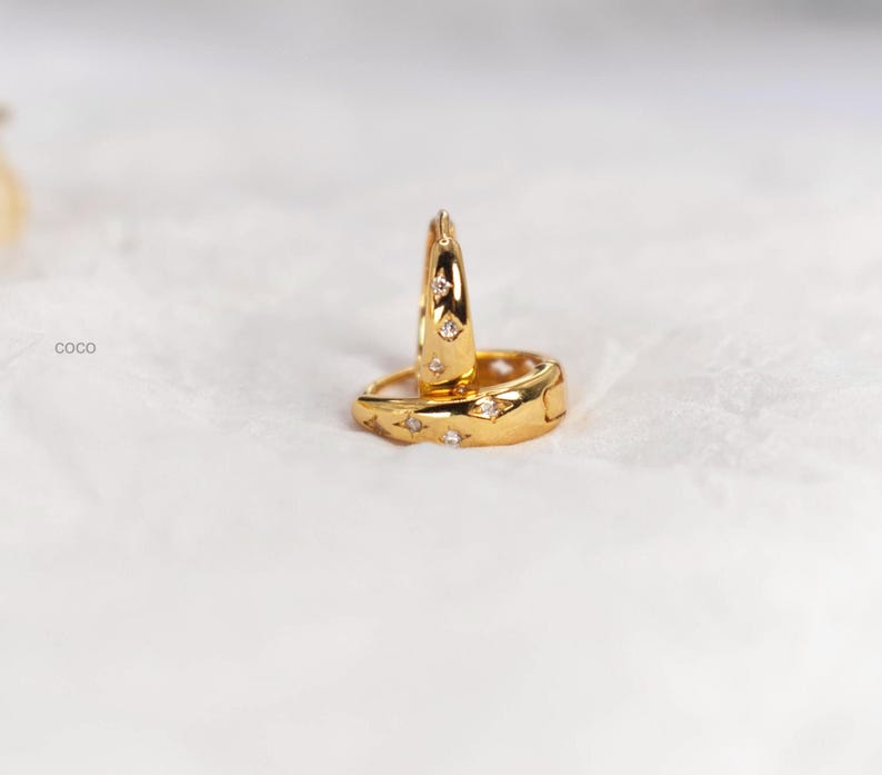 May include: A pair of gold hoop earrings featuring a polished finish and small, star-shaped engravings with embedded clear stones. The word coco is printed in small black letters to the left of the jewellery. The hoops have a hinged closure.