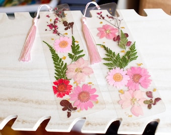 Puede incluir: Dos marcapáginas de flores prensadas rosas y verdes con borlas rosas. Los marcapáginas están hechos de plástico transparente y tienen una variedad de flores y hojas prensadas.