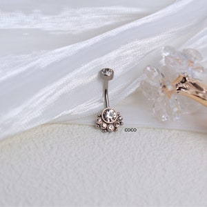 May include: A silver belly button ring with a clear crystal center and a row of smaller crystals around it. The ring is on a white fabric background.