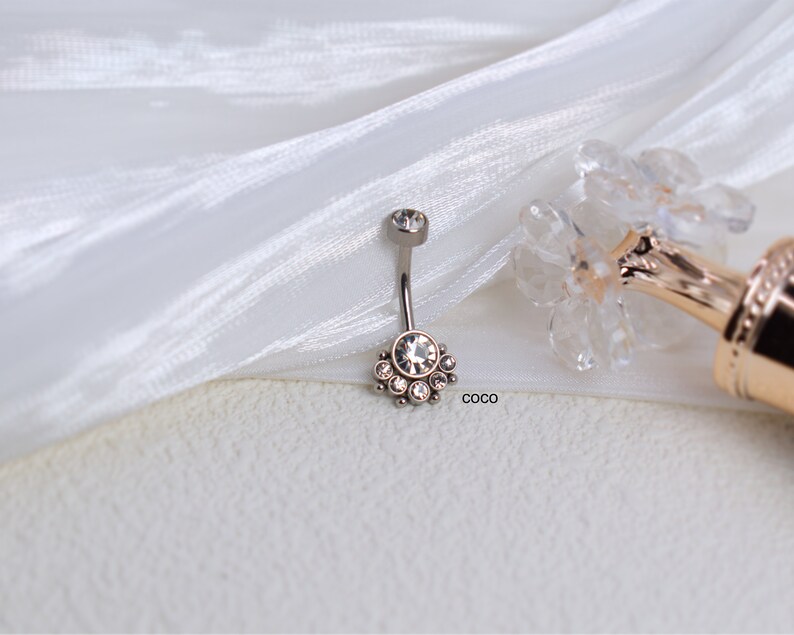 May include: A silver belly button ring with a clear crystal center and a row of smaller crystals around it. The ring is on a white fabric background.