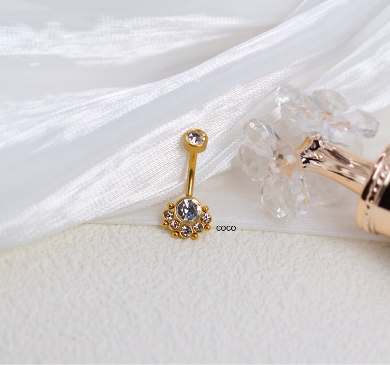 May include: Gold belly button ring with a clear crystal center and a circle of smaller clear crystals around it.  The ring is on a white fabric background.  The text "COCO" is visible in the lower right corner of the image.