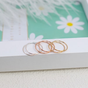 May include: A set of three thin, circular hoop rings in silver, rose gold, and yellow gold tones. Each ring features a simple, continuous band design with a small opening, resting on a white surface.