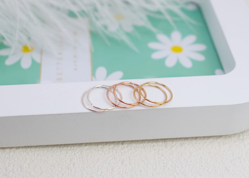 May include: A set of three thin, circular hoop rings in silver, rose gold, and yellow gold tones. Each ring features a simple, continuous band design with a small opening, resting on a white surface.