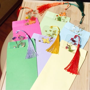 May include: Seven colorful envelopes with pressed flower bookmarks inside. Each bookmark has a different flower design and a tassel.