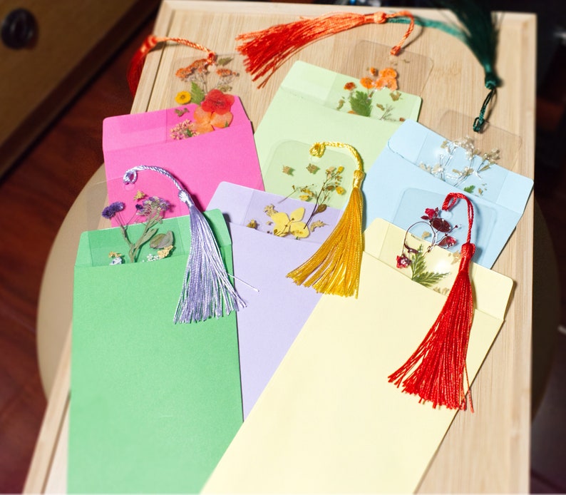 May include: Seven colorful envelopes with pressed flower bookmarks inside. Each bookmark has a different flower design and a tassel.