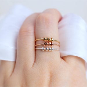 May include: Three delicate rings in gold, rose gold, and silver, each featuring three small, evenly spaced beads. The rings are displayed on a hand, showcasing their minimalist design and varying metallic finishes. The background is a soft, white fabric.